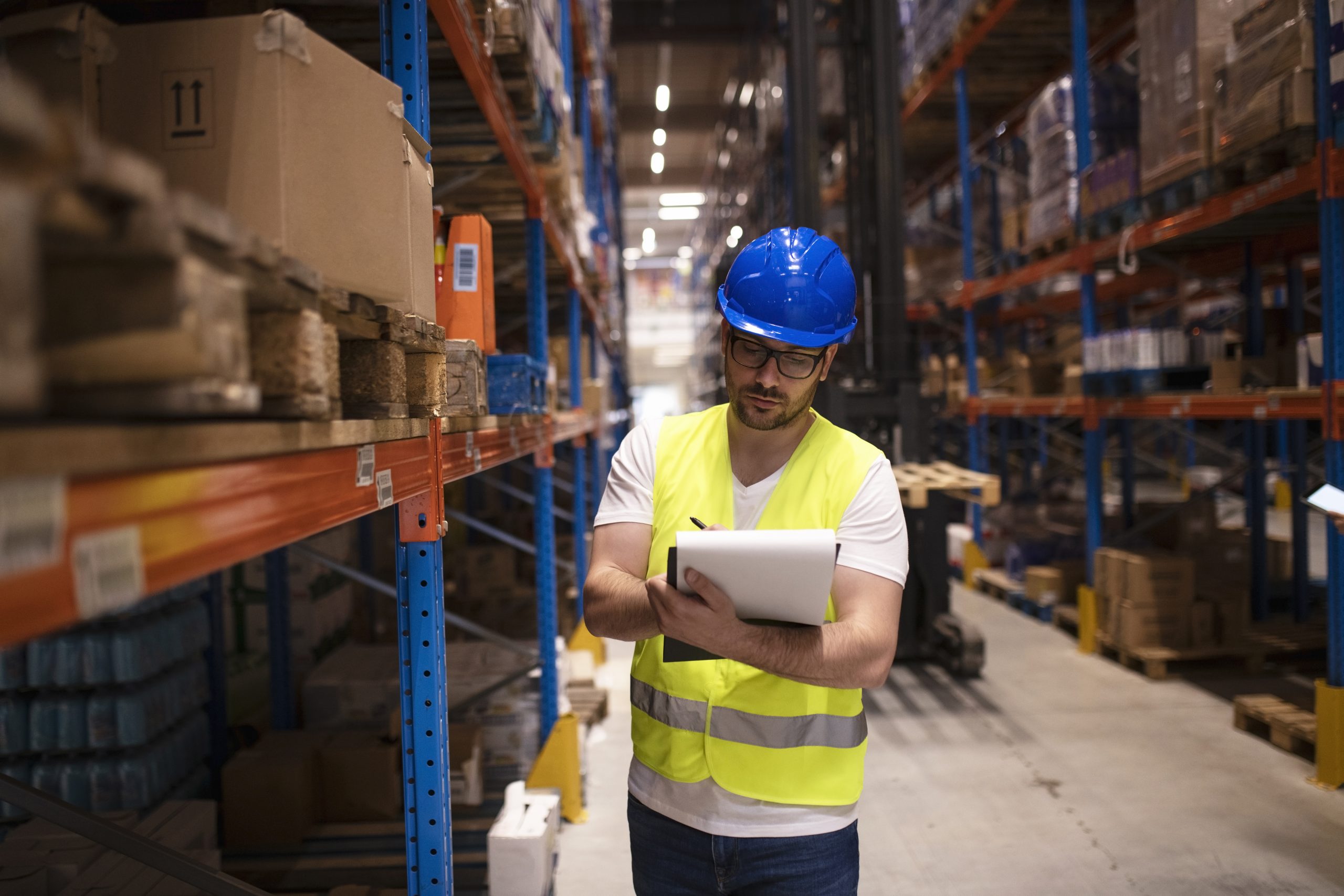 man with a blue helmet writing notes in a big warehouse 